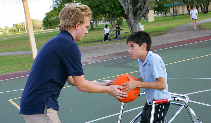 Typically-abled boy passes basketball to special needs boy in a walker on a basketball court