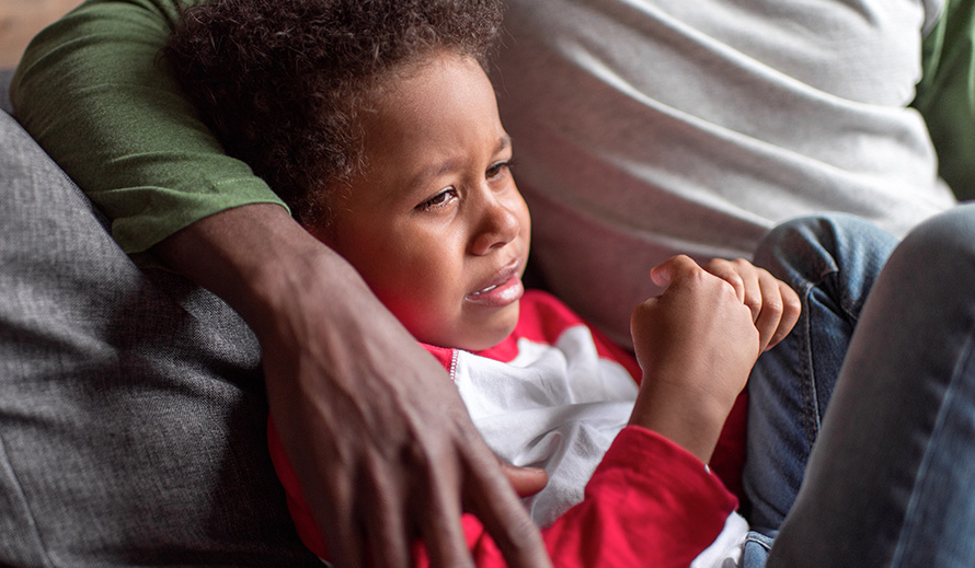 A distressed young African-American boy curled up on the couch being comforted by his father.