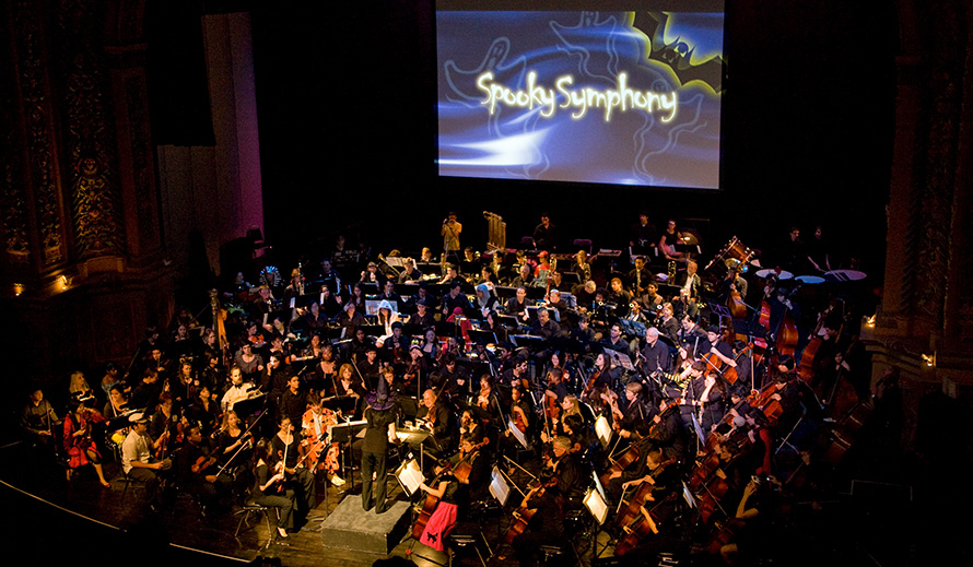 A costumed symphony orchestra performing a concert while cartoon ghosts, bats and the words “Spooky Symphony” are projected onto a big screen above them.