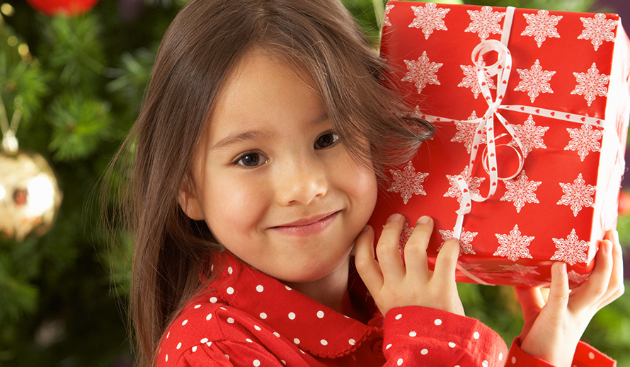 Smiling pajama-clad little girl holding a wrapped gift in front of a Christmas tree