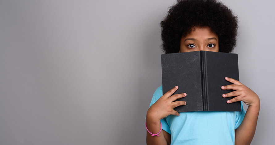 A shy African American girl hides behind a book. 