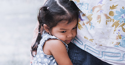 A little Hispanic girl, crying and standing next to her mother with her arms wrapped around her waist.